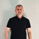 Adult male posing casually with a smile against a plain backdrop, wearing a black shirt and jeans.